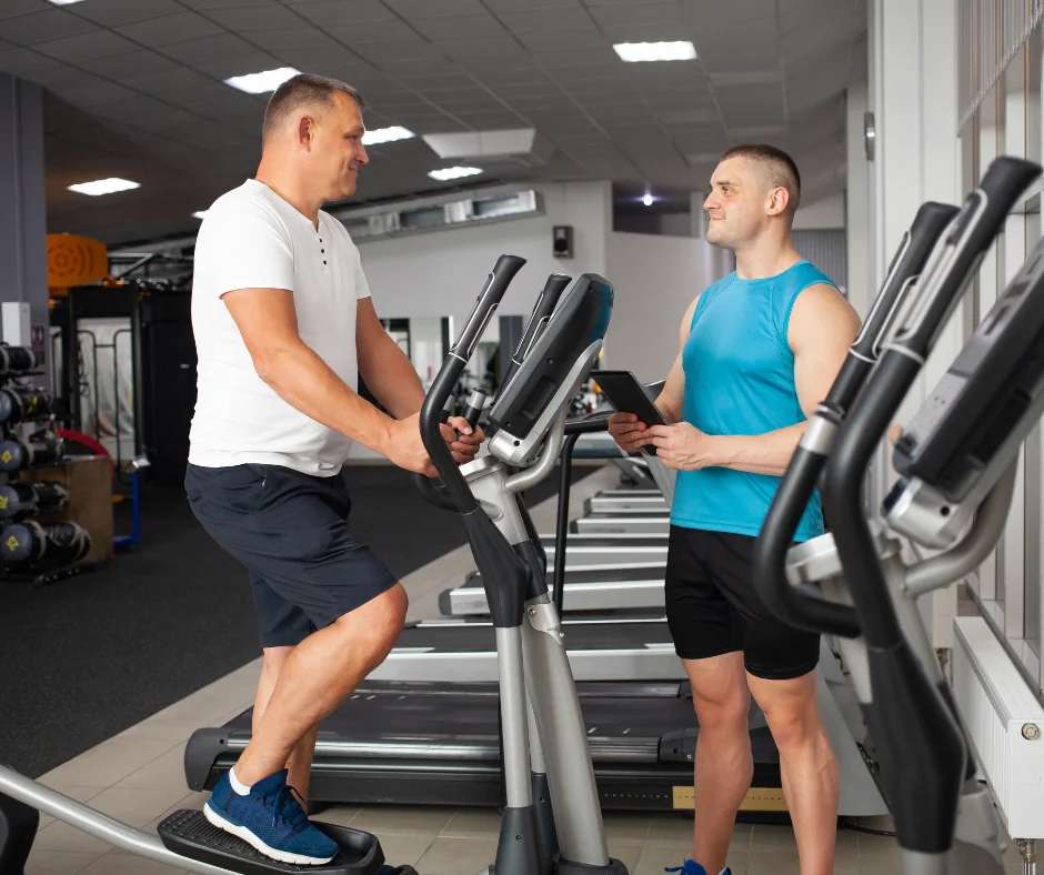 An exercise physiologist holding a tablet while monitoring a man training on an elliptical cross-trainer in a gym.