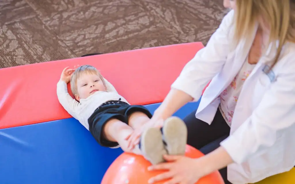 A paediatric exercise physiologist guiding a young boy through leg elevation and mobility exercises on a red exercise ball.