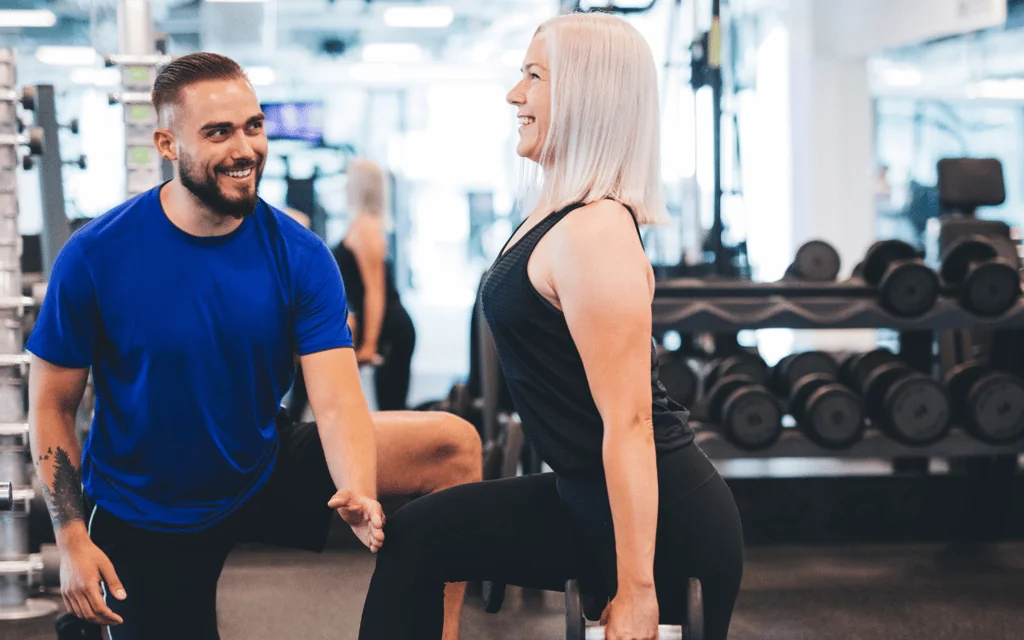 An exercise physiologist in a gym setting guiding a woman through a dumbbell lunge during an in-person training session.