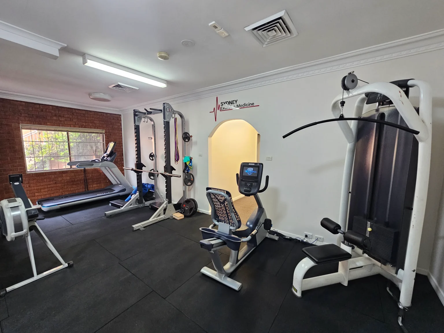 An interior view of a fully equipped exercise physiology clinic gym with a treadmill, squat rack, stationary bike, and lat pulldown machine.