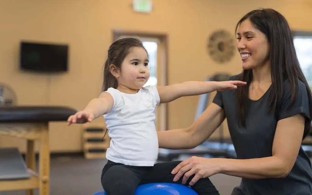 A female exercise physiologist helping a young girl balance on a therapy ball with arms extended.