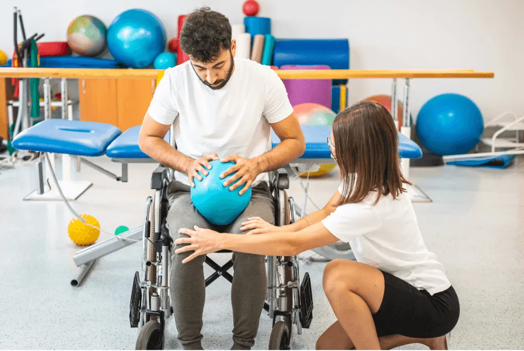 An exercise physiologist guiding a man in a wheelchair during a therapy session with a small exercise ball.