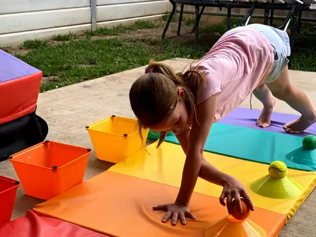 A young girl doing a downward dog position while picking up balls on a colorful exercise mat during a physical therapy session.