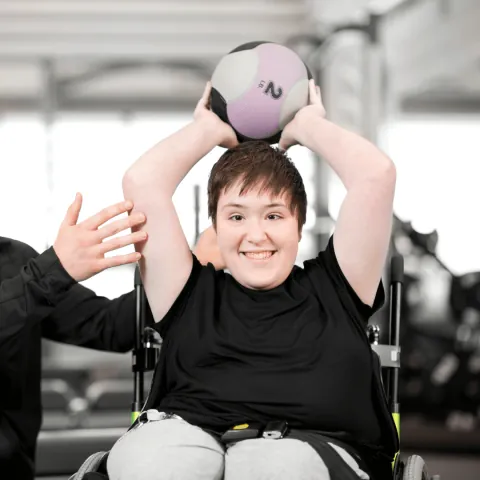 A smiling person in a wheelchair doing an overhead medicine ball exercise with a trainer assisting.