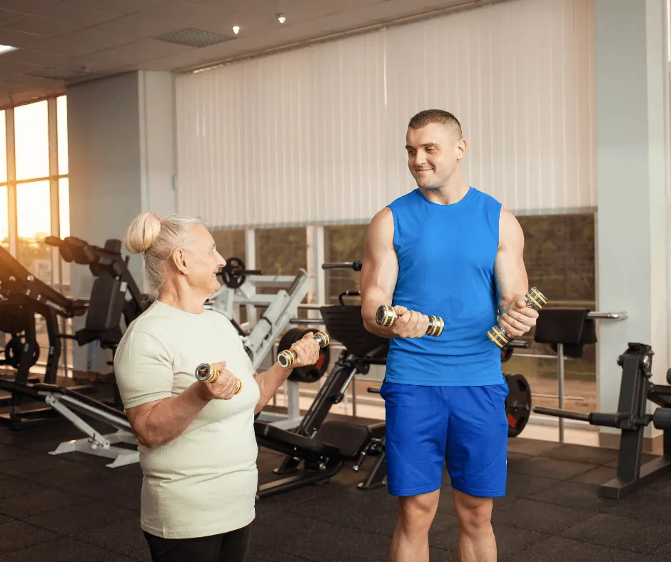 An exercise physiologist guiding an elderly woman doing dumbbell bicep curls in a gym setting.