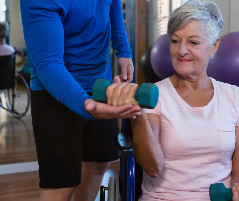 An exercise physiologist supporting a senior woman lifting a dumbbell during a rehabilitation session.