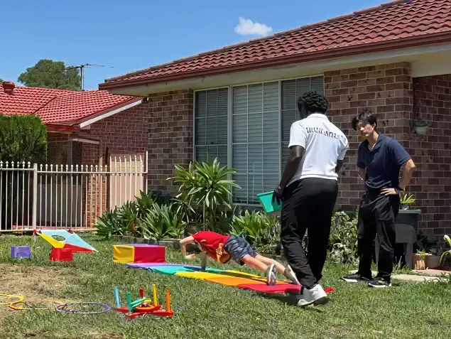 A child on a rainbow mat doing exercises in a Sydney backyard with an NDIS mobile exercise physiologist, while a second supervisor watches.