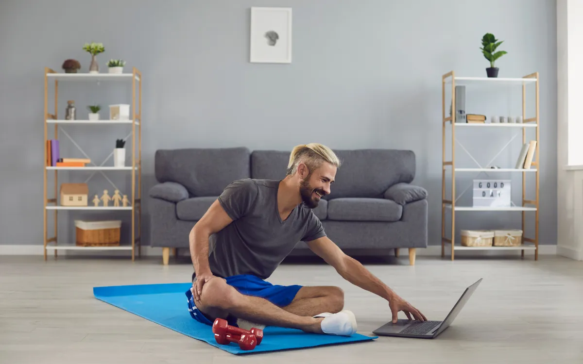 A man sitting on an exercise mat at home using his laptop for an online telehealth workout session.