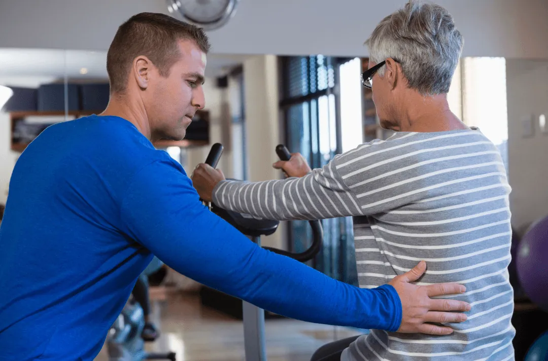 An accredited exercise physiologist guiding an older woman on a stationary exercise bike during a rehab session.
