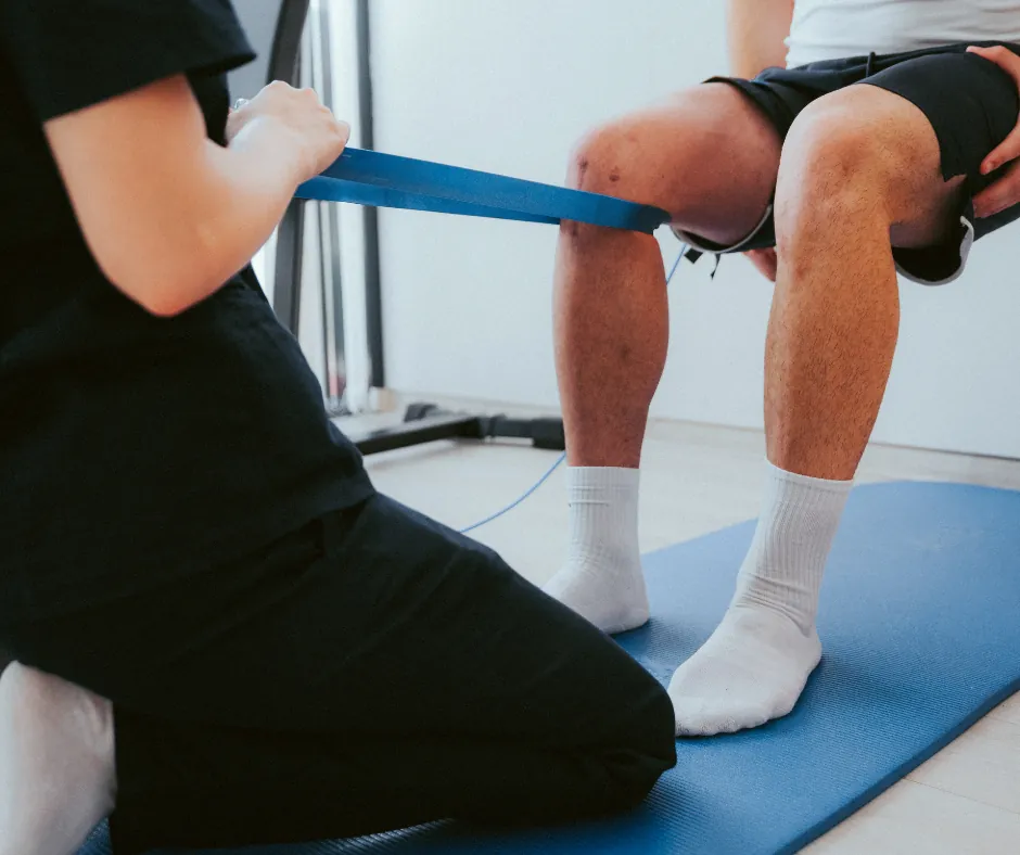 An exercise physiologist assisting a patient with a blue resistance band around the knees during a rehabilitation exercise.